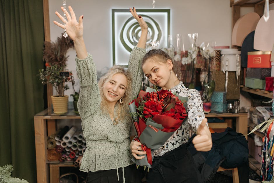 Two young women inside a floral shop, with one holding a bouquet of vibrant red roses wrapped in grey and red paper, and the other raising her hands in celebration. The woman with the roses is dressed
