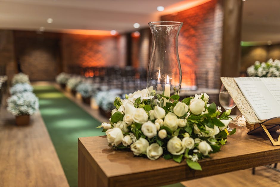 A floral arrangement featuring a large bouquet of fresh white roses with lush green foliage, positioned on a wooden table alongside a glass hurricane lantern and an open book of sheet music, set in an