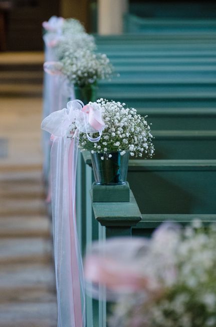 A row of small floral arrangements placed in silver metal buckets on a dark green wooden pew at a wedding venue, decorated with pink and white striped ribbons flowing down the side. Each arrangement f