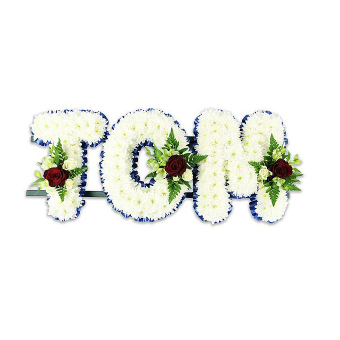 Presented on a clean white field, the name TOM is rendered in large sculptural letters entirely covered in countless small, immaculate white chrysanthemums, their rounded petals packed closely to form a soft, pillowy surface that reads as both purity and presence. A vivid royal blue ribbon outlines each letter with a crisp, graphic line, the colour adding refined contrast and a gentle formality that helps the letters sit clearly in a chapel or funeral hall. Each letter is accented with a compact floral spray centred on a single, deep red rose-its velvety throat a deliberate emblem of love and respect-accompanied by sprightly cream spray roses and fine fern fronds that provide lift and natural texture against the chrysanthemum field. The construction is supported by a discreet dark metal bar at the back and base, ensuring the letters can be displayed upright on an easel or floor stand without sagging. Under soft, neutral lighting the white chrysanthemums read as luminous, the blue ribbon a neat frame and the red roses a poignant visual anchor; the scent would be quietly floral, a restrained blend of rose perfume and fresh greens. This is a craft-led memorial piece, made by experienced hands with attention to balance, volume and the quiet language of flowers. For families in Esher - whether gathering near Esher Green, by Claremont Landscape Garden, or at a local chapel - this personalised tribute offers a composed, heartfelt way to commemorate a loved one, designed to sit respectfully within ceremonial spaces.