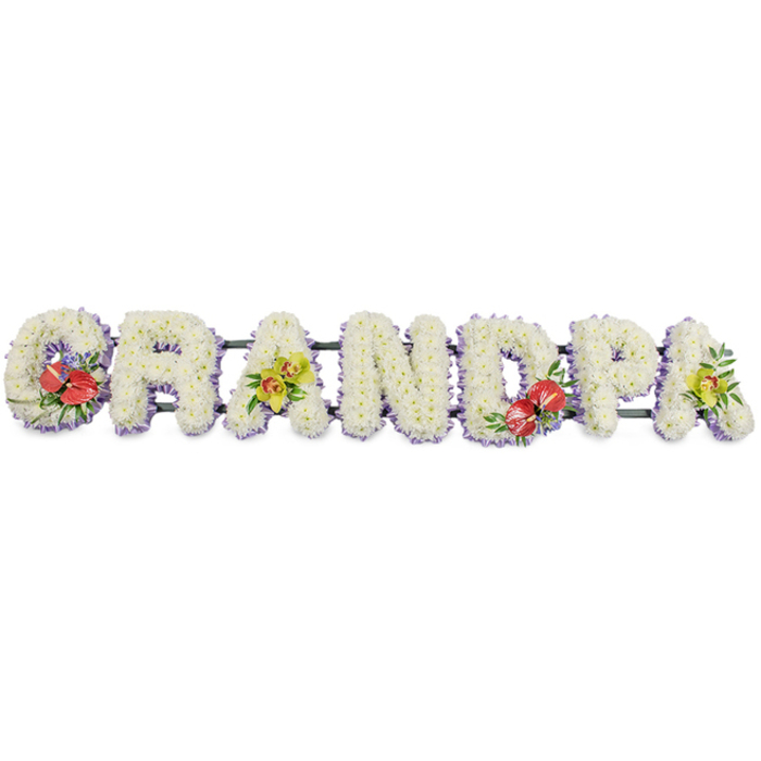 This photograph shows a striking floral tribute spelling out GRANDPA in large, block capital letters, a design that feels both traditional and personal, as if made to rest at a funeral service in Esher. Each letter is fully clothed in white chrysanthemums, their densely packed blooms forming a soft, cushion-like surface that suggests purity, comfort, and the gentle closeness of family. The chrysanthemum petals overlap in layers, catching the light delicately and adding a sense of depth and texture to the otherwise simple white field. A slim edging of pale lavender ribbon runs around the base of each letter, its muted purple tone subtly defining the shapes and introducing a hint of colour that feels calm and meditative. The letters are aligned on deep green support bars that run behind them from left to right, ensuring the tribute remains strong and stable for display on stands or within a hearse. Carefully placed accent sprays of flowers break up the expanse of white: on the 'G' and the 'D', clusters of deep red anthuriums emerge, their glossy, heart-shaped spathes and bright yellow spadices rising from a nest of dark green foliage and tiny filler blooms. On the first 'A' and the last 'A', yellow-green orchids are arranged in graceful arcs, their refined, starry blooms adding elegance and a slightly modern feel. The whole composition evokes the quiet surroundings of Esher's leafy streets and green spaces, such as nearby Esher Common, where families might reflect after a service. It is easy to picture grandchildren and relatives choosing this piece as a final, heartfelt message to a beloved grandfather, the flowers speaking gently of love, respect, and treasured memories.