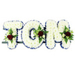 The image presents a traditional funeral flower tribute spelling out the name TOM in three bold, three-dimensional letters, arranged side by side on a slim dark frame. Each letter is meticulously filled with small white chrysanthemums, the blooms packed closely together to form a smooth, cloud-like surface with a soft, comforting appearance. The white petals create a sense of purity and stillness, ideal for a moment of remembrance. Around the edge of each letter runs a royal blue satin ribbon, gently pleated so its folds catch the light, giving a subtle shimmer that crisply outlines the shapes and adds a dignified accent of colour. On the surface of the 'T', 'O', and 'M', carefully placed floral clusters bring richness and depth: velvety deep red roses, delicate white or cream spray roses, and slender white flowers reminiscent of freesia are interwoven with glossy green foliage and feathery fern fronds. These sprays sit like small bouquets, softening the block capitals and drawing attention to the name. The tribute is photographed against a clean white background, allowing every tone and detail to stand out clearly-the interplay of white, blue, red, and green feels balanced and heartfelt. It is the type of personalised arrangement that might be chosen by a family in Esher for a funeral at a nearby church or crematorium, perhaps after being prepared by an experienced florist familiar with local services. The overall impression is one of quiet dignity and deep affection, a floral memorial created to honour and remember a loved one named Tom with grace and care.